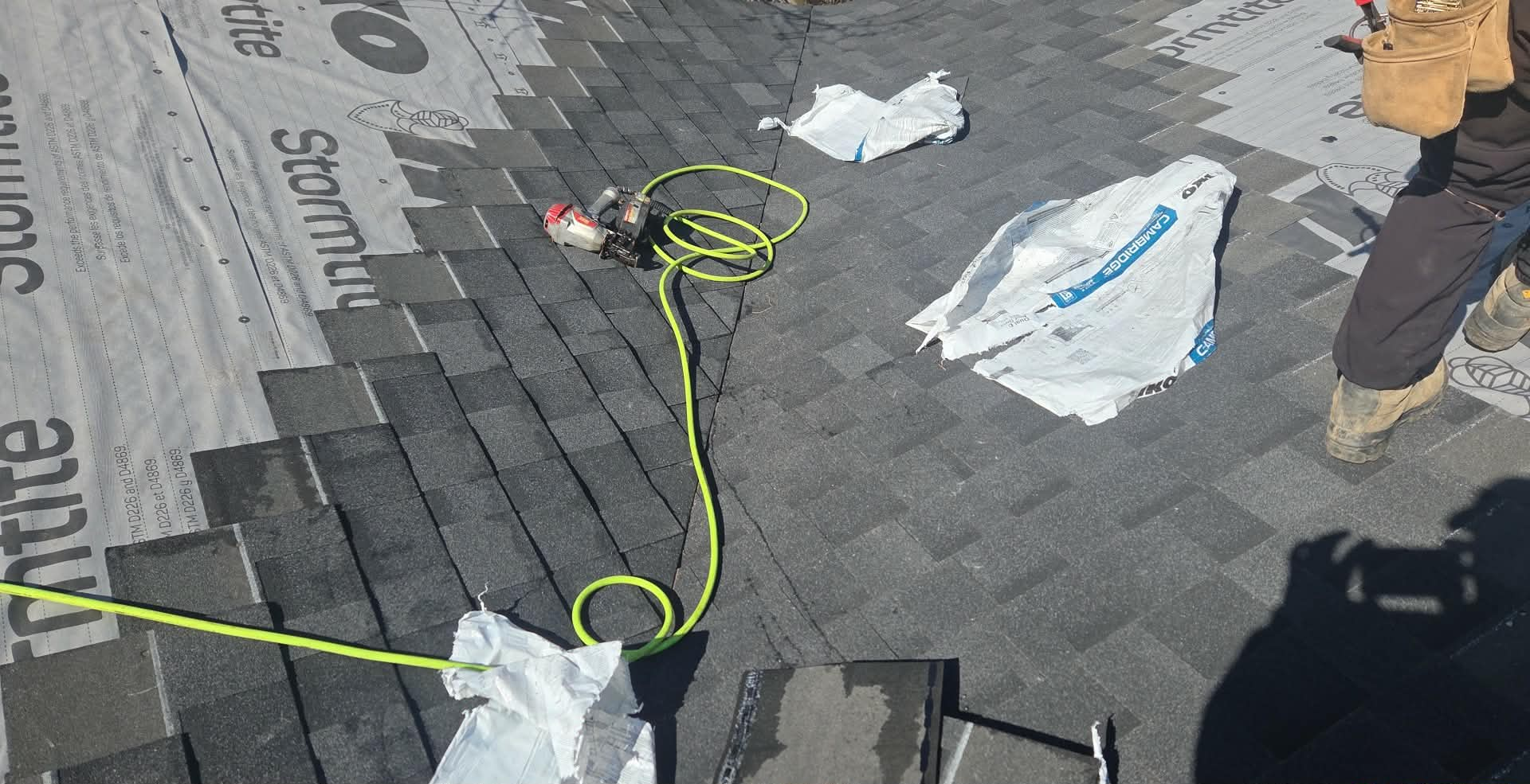 Roof repair tools and debris on gray shingles, with a neon green cord and workers’ boots visible
