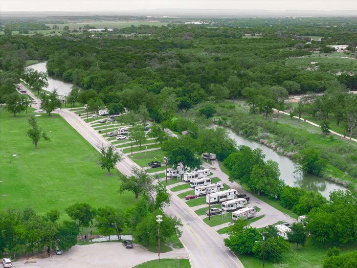 Aerial view of a park with green fields, trees, a winding river, and parked cars along a road