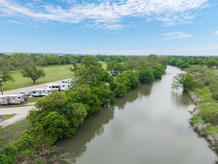 Aerial view of a river bordered by lush green trees, with a row of white RVs parked along the left bank.
