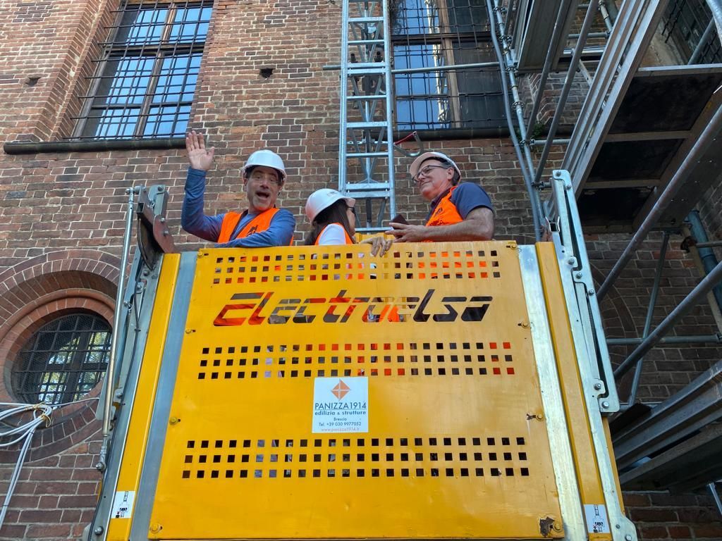 Three people in safety vests on a yellow Electrolsa lift, waving in front of a brick building with scaffolding.