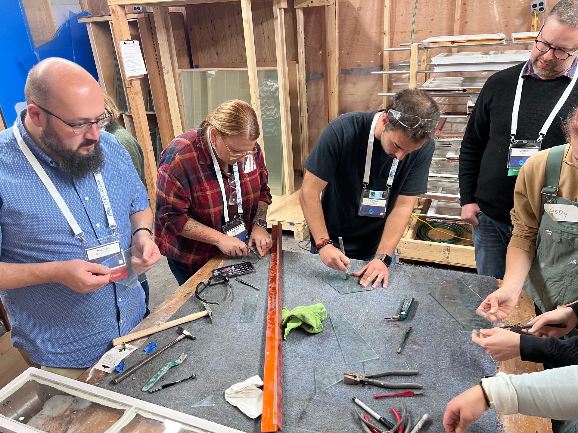 People working on a table with tools, possibly stained glass. Workshop setting with wood framing.