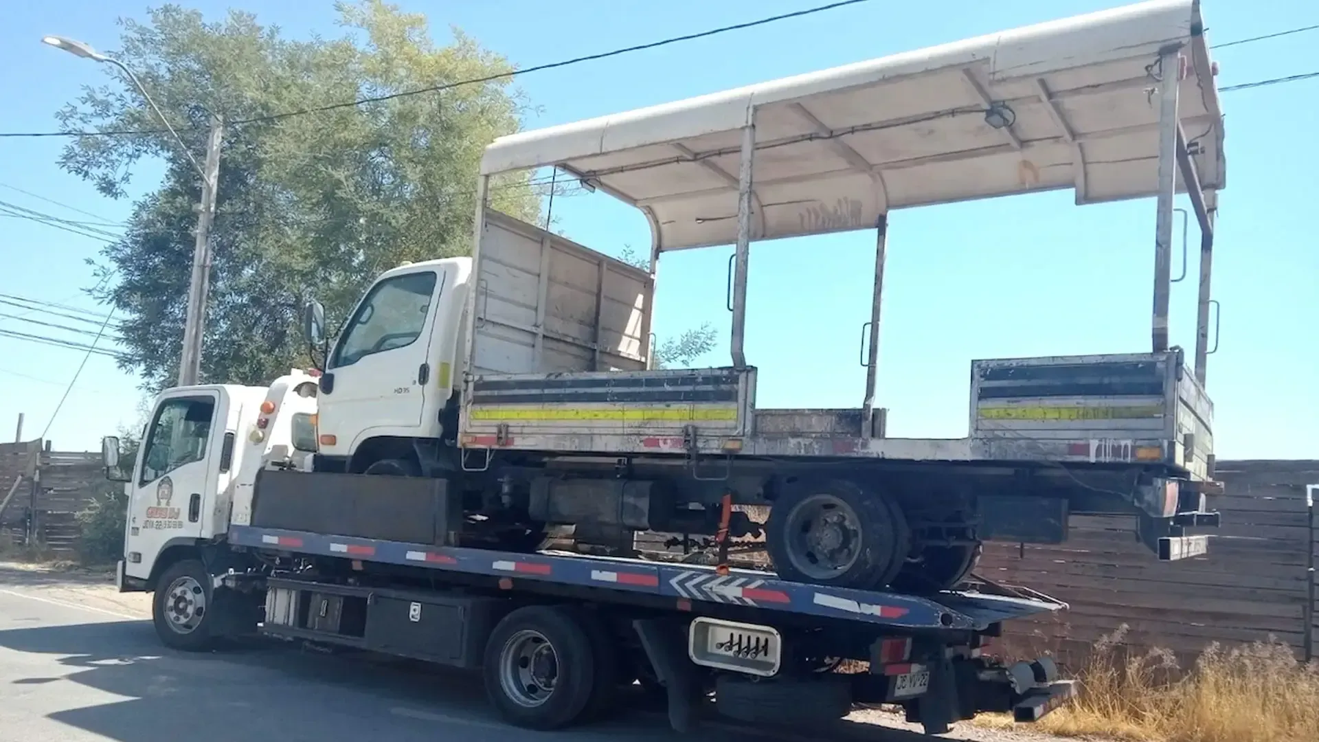 Una grúa de plataforma que transporta un camión de plataforma blanco con una estructura metálica tipo toldo en una carretera pavimentada y soleada.