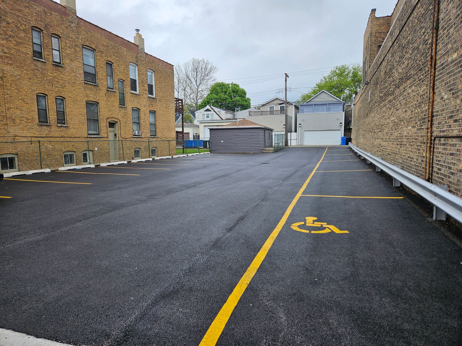 Empty asphalt parking lot with yellow lines beside brick buildings and a metal fence