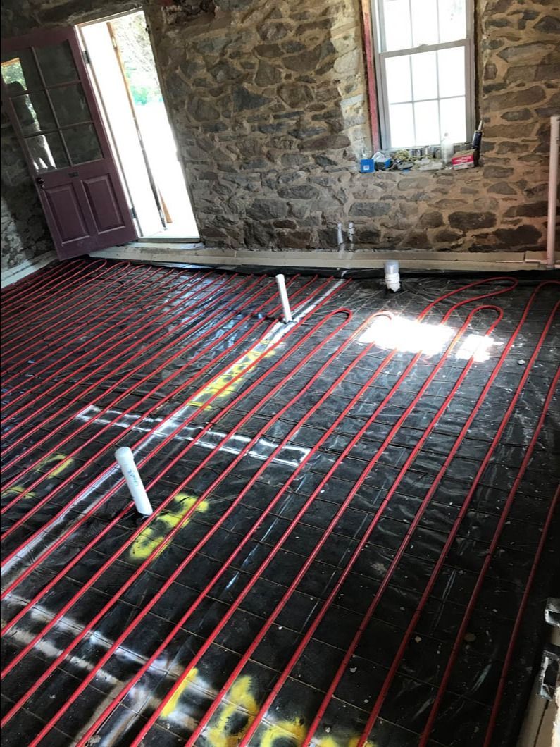 Stone room floor under installation, with black underlayment, red heating tubes, and white pipes near a door and window