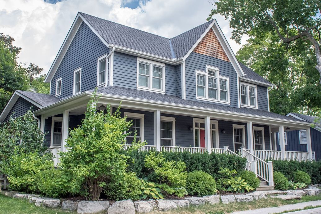 Blue-gray house with white trim and a wraparound porch, surrounded by lush green shrubs and trees