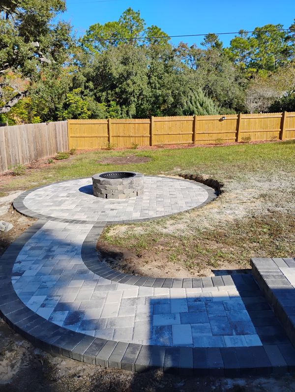 A stone fire pit sits in the center of a circular paver patio, connected by a curved walkway to an adjacent paved area. A stone fire pit sits in the center of a circular paver patio, connected by a curved walkway to an adjacent paved area.