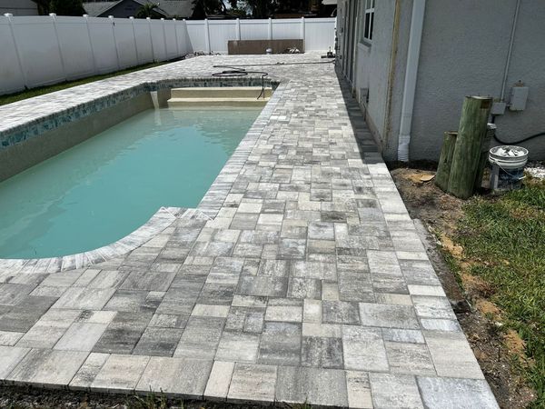 A rectangular swimming pool surrounded by a light-colored, multi-sized paver patio, with a white vinyl fence in the back. A rectangular swimming pool surrounded by a light-colored, multi-sized paver patio, with a white vinyl fence in the back.