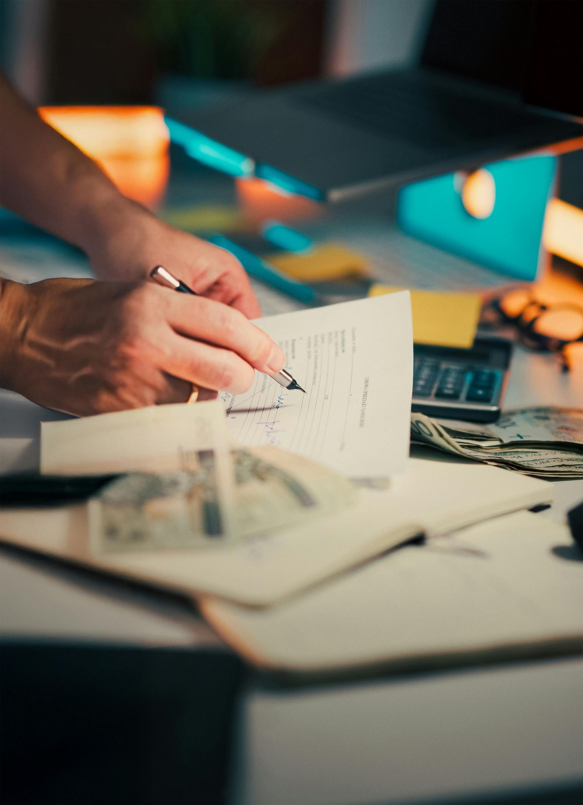 Hands arranging papers on a desk beside a lit monitor and scattered documents