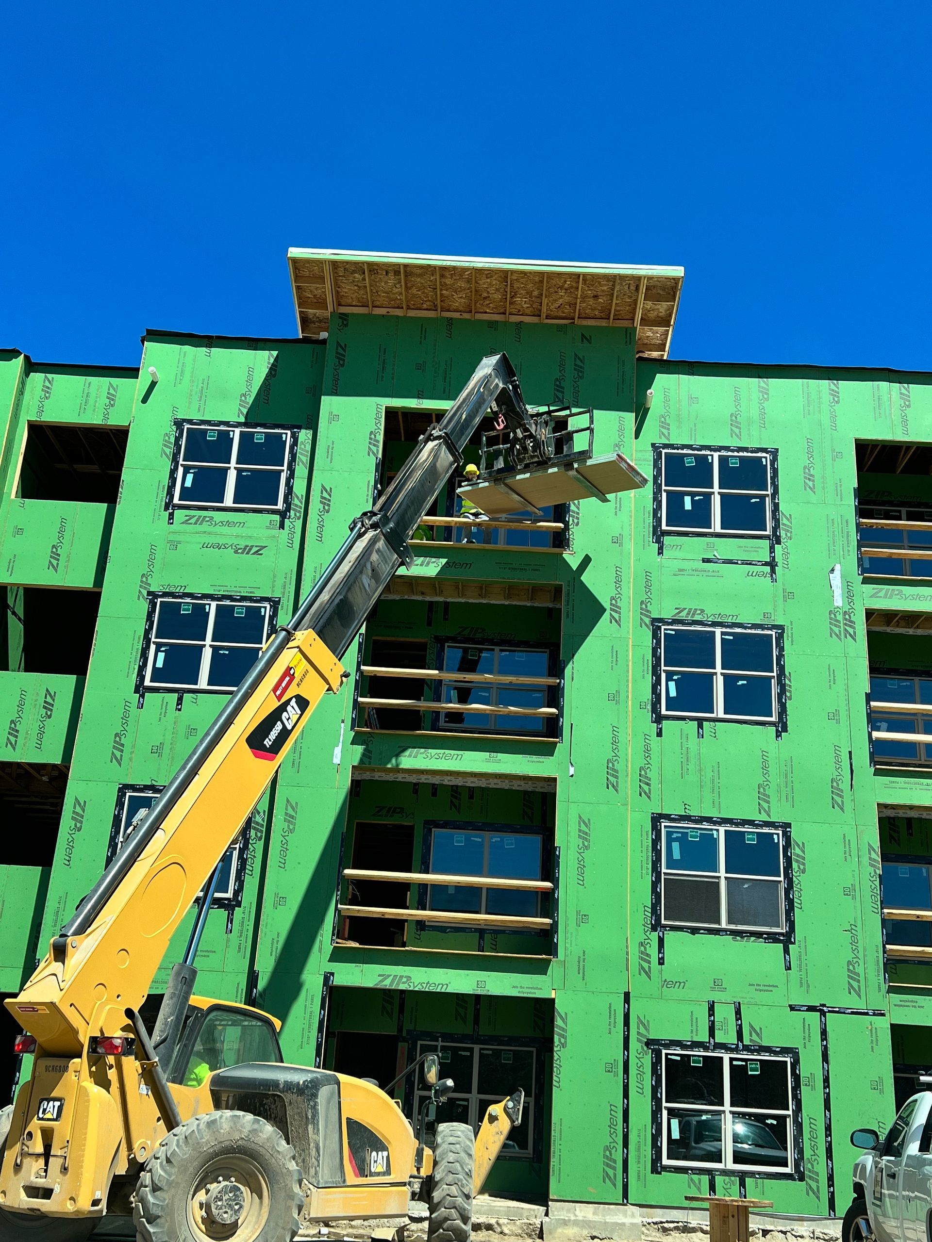 A yellow forklift is lifting a piece of wood in front of a green building under construction.