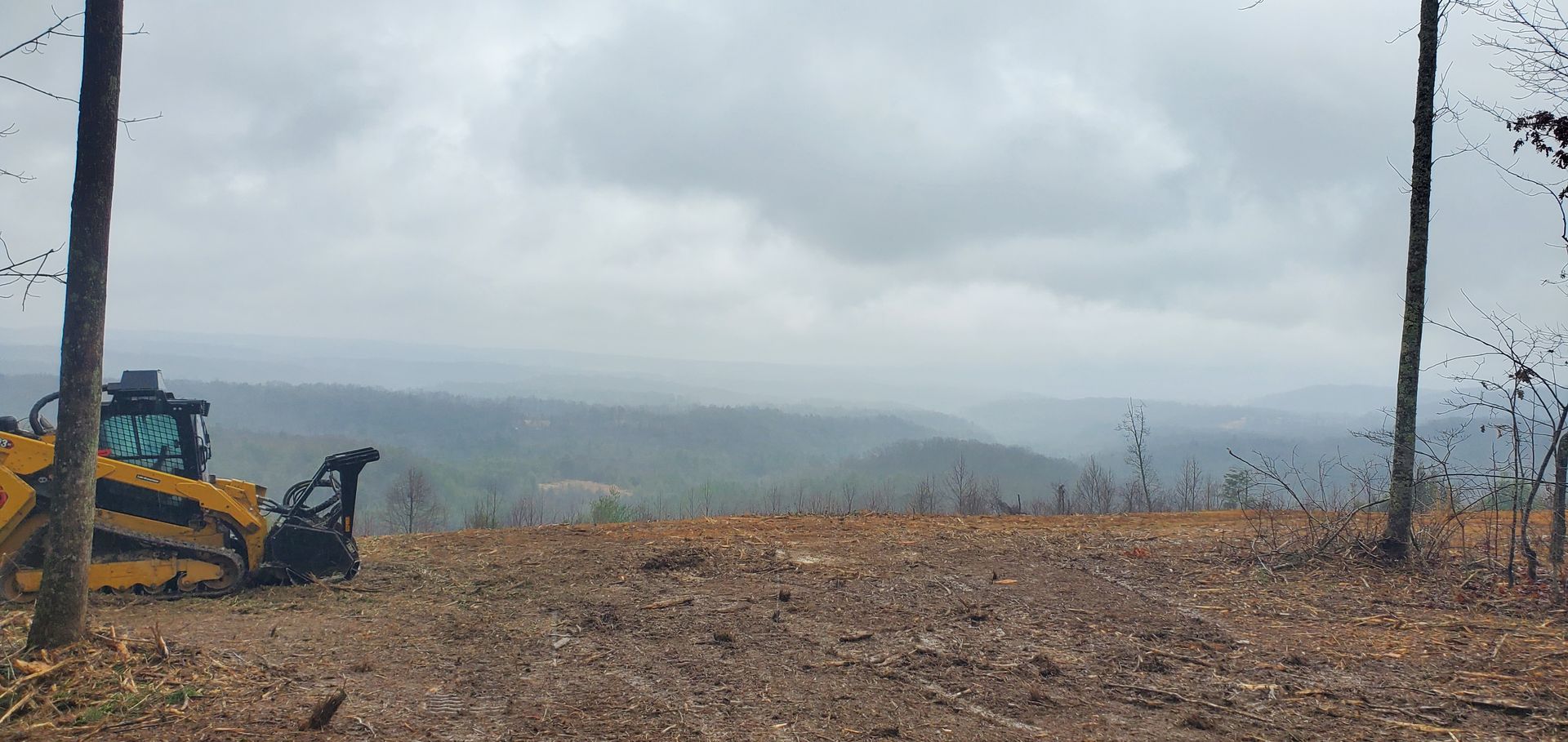 A yellow construction vehicle sits on a cleared, brown hillside overlooking a vast, foggy mountain valley.