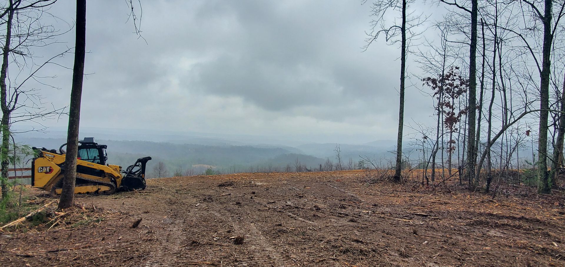 A yellow bulldozer parked on a cleared, muddy hillside overlooking a wide, hazy valley under a cloudy sky.