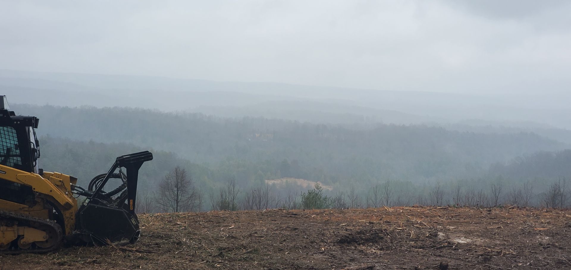 A yellow construction vehicle sits on a dirt ridge overlooking a vast, foggy forest valley under a cloudy sky.
