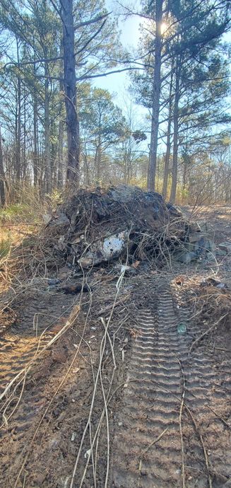 A large pile of dirt, brush, and debris sits in a wooded area with visible bulldozer or tractor tread marks in the mud.