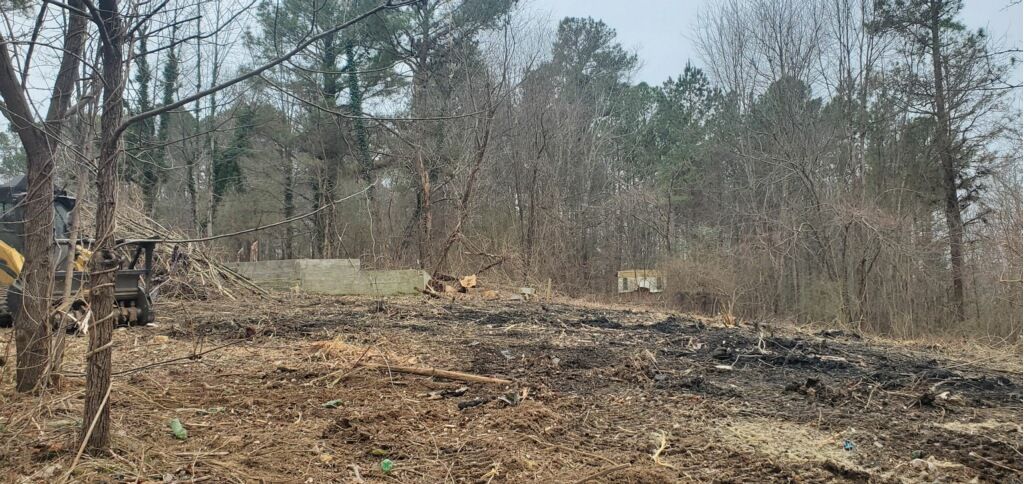 A cleared, scorched patch of earth with a bulldozer visible on the left and a small building in the wooded background.