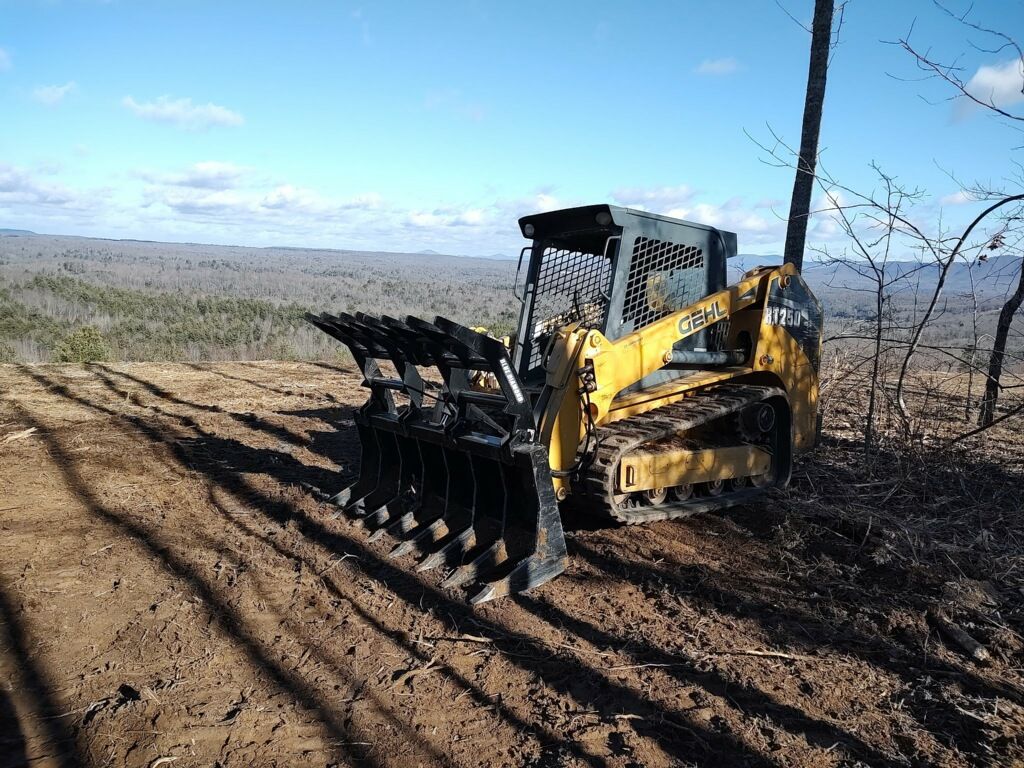 A yellow compact track loader with a root rake attachment parked on a cleared, hilly landscape under a blue sky.