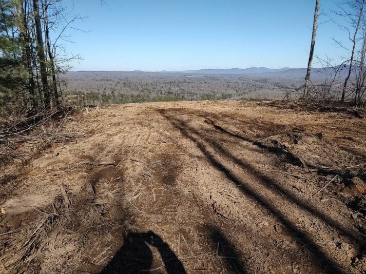 A wide, barren patch of cleared land with tire tracks overlooking a forested valley under a clear blue sky.