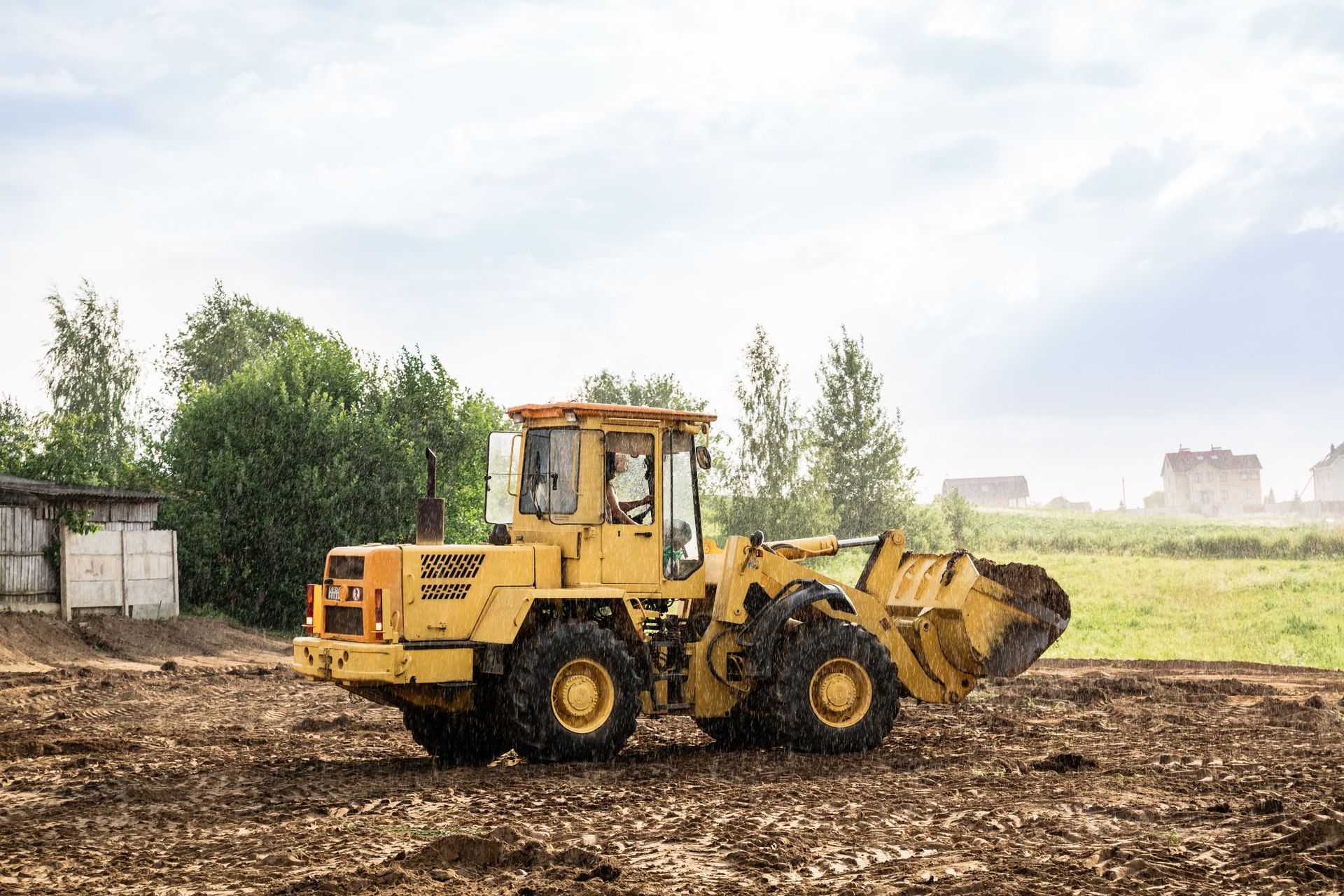 A yellow front-end loader parked on a dirt field with trees and a building in the background under a bright, cloudy sky.