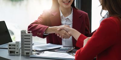 Two businesspeople shaking hands over a desk with building models and documents in a bright office