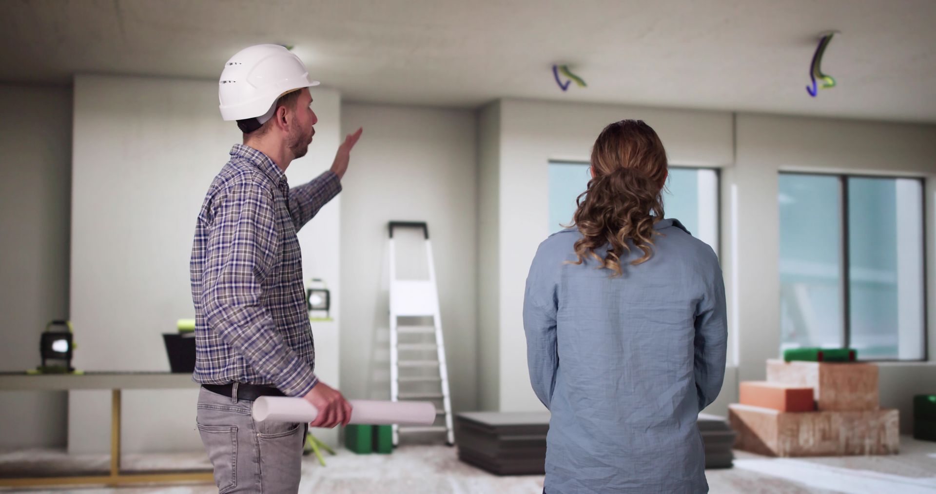 Two people in a partially finished room, one in a hard hat gesturing toward the wall