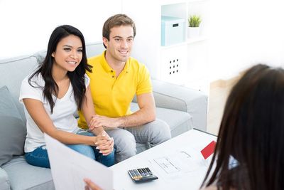 Two people sit on a couch speaking with a person across a coffee table in a bright living room.