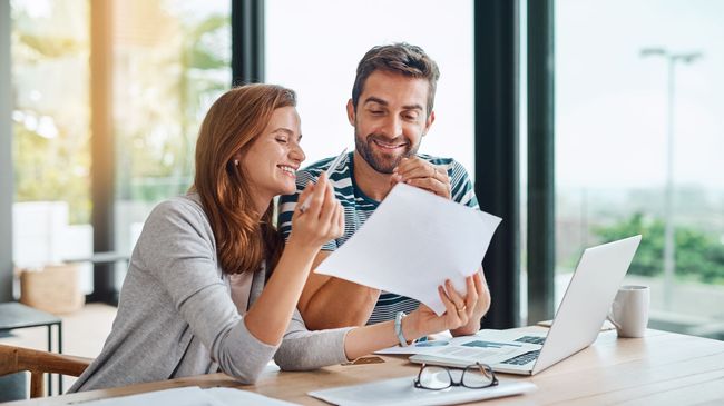 Two coworkers smiling and reviewing a document at a desk with a laptop in a bright office
