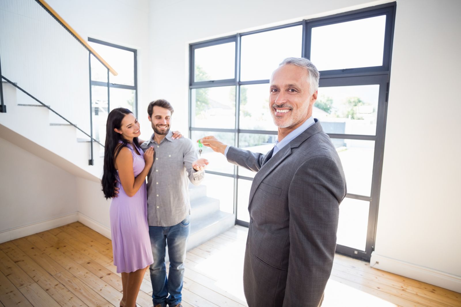 Three people in a bright room, smiling and pointing toward large windows.