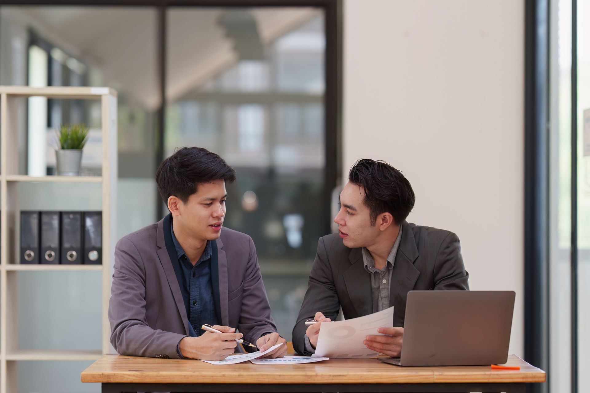 Two coworkers talking at a desk with papers and a laptop in a bright office