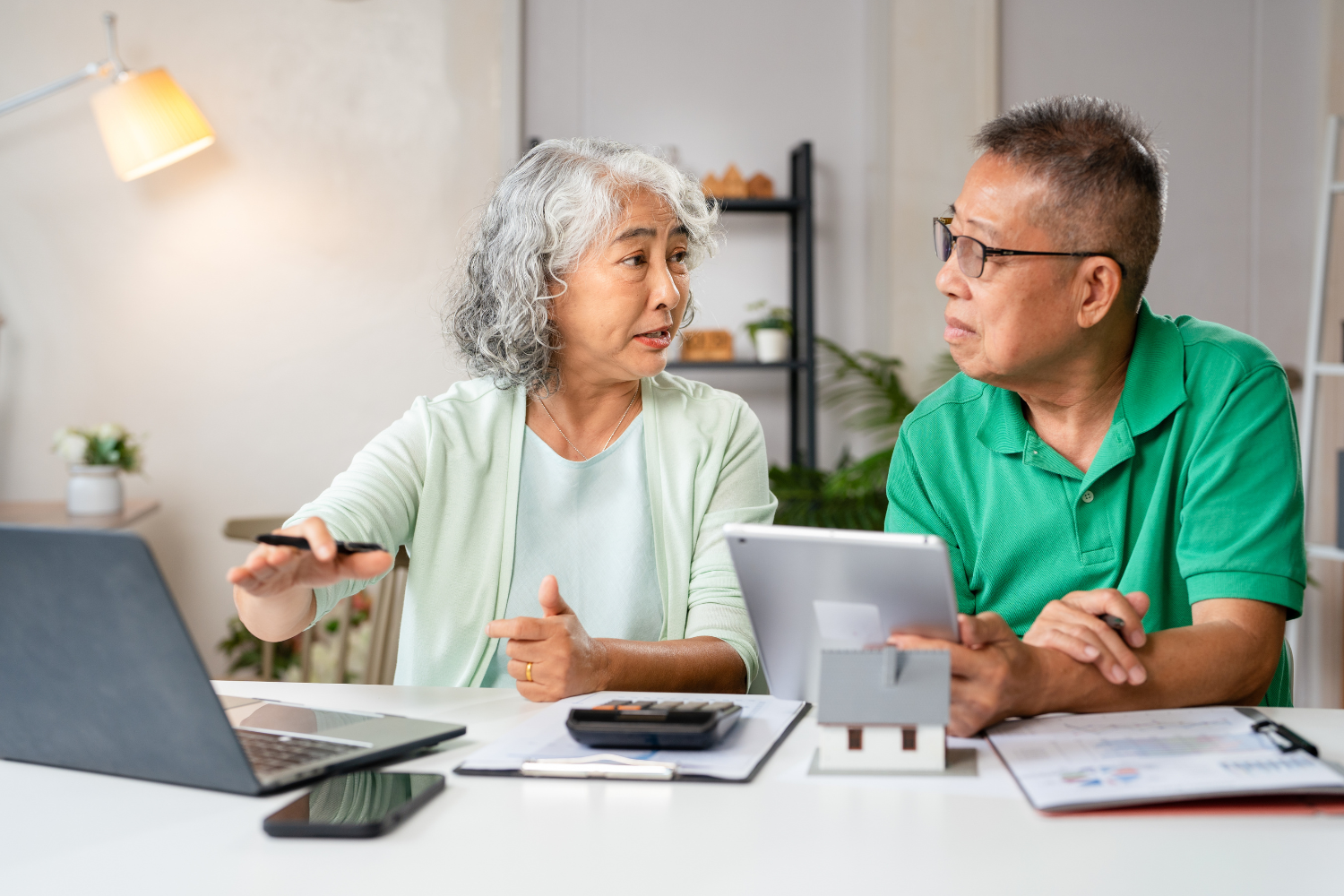 Elderly Asian couple reviewing finances together at a table, using laptop, tablet, and calculator.