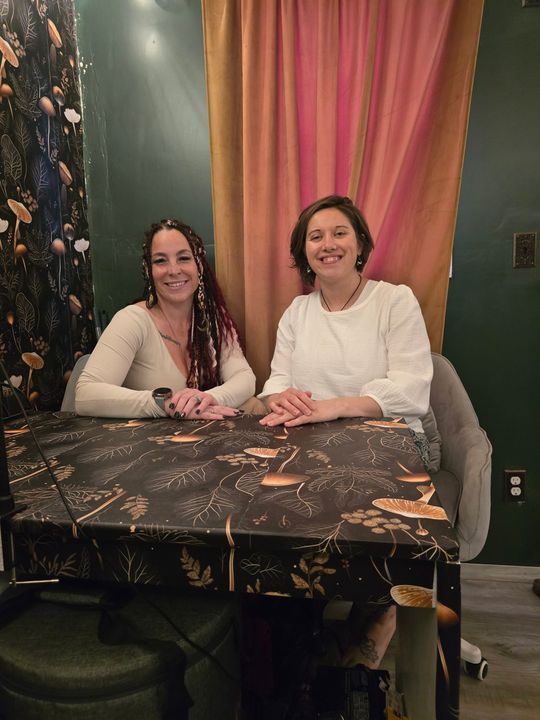 Two women sitting at a desk in front of a gold window curtain. Two women sitting at a desk in front of a gold window curtain.