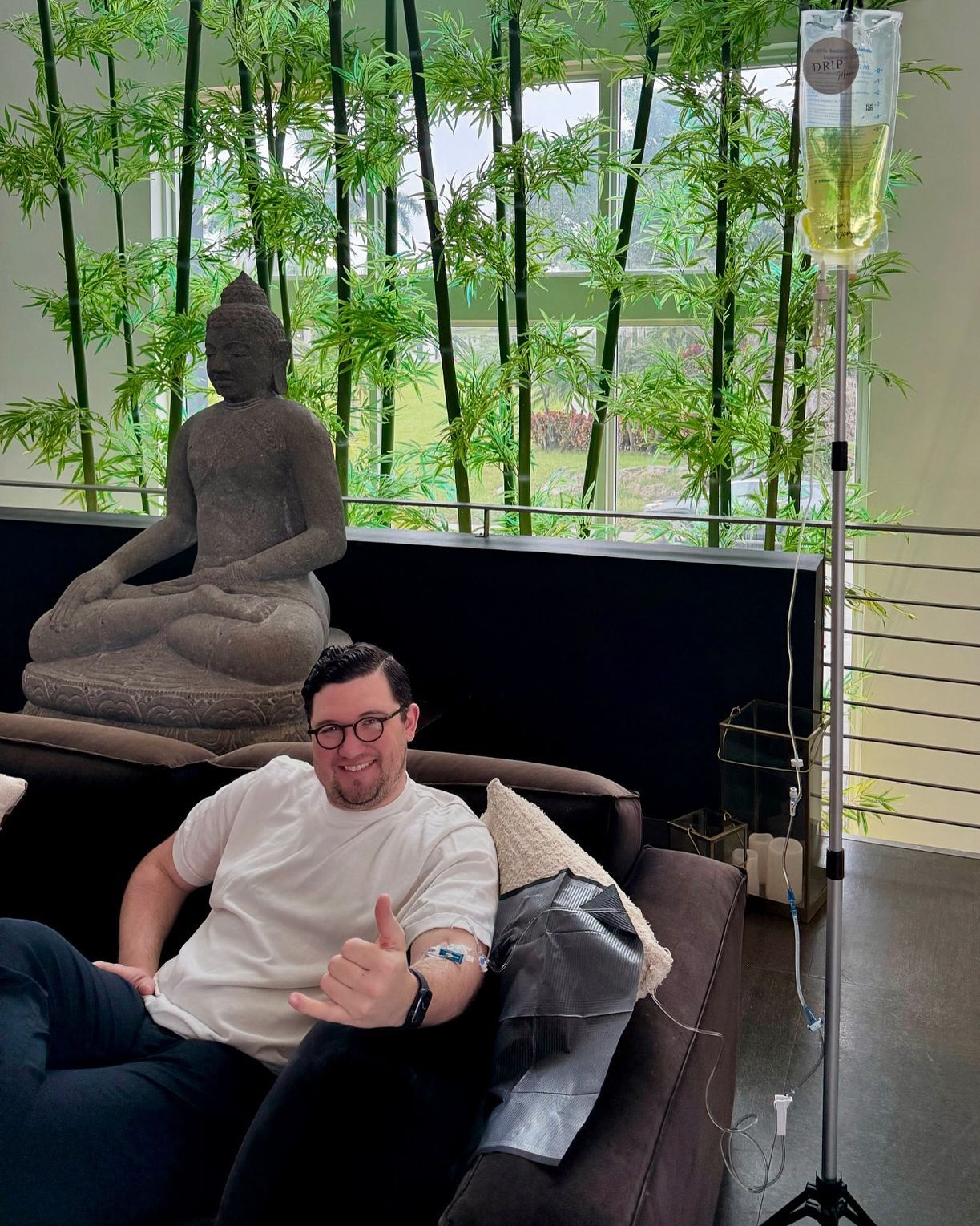Man receiving IV drip, reclined on couch. Buddha statue, bamboo backdrop. Smiling, giving a thumbs-up.