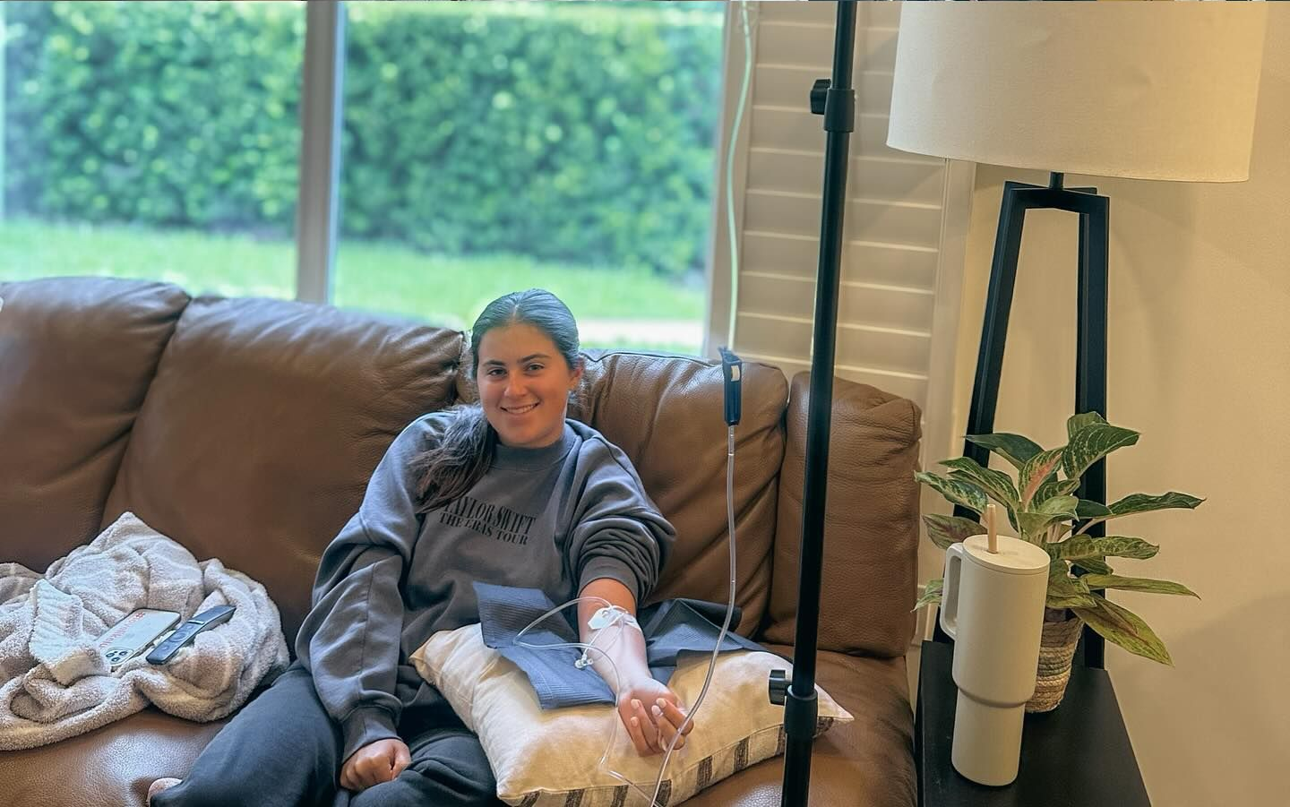 Woman on couch receiving an IV, smiling. In a living room, near a lamp and plant.