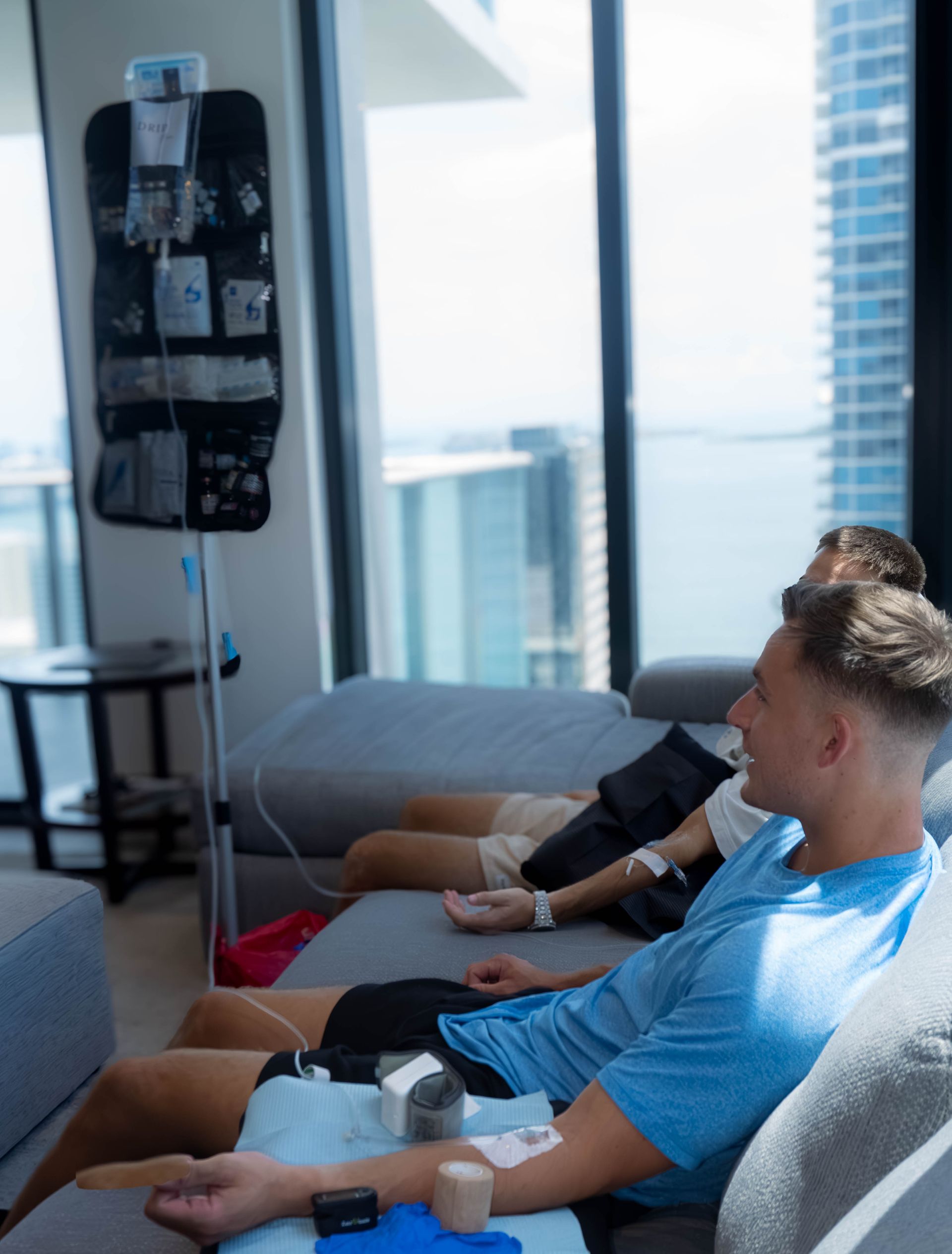 Two people receive IV drips on couches in a modern room with a city and water view.