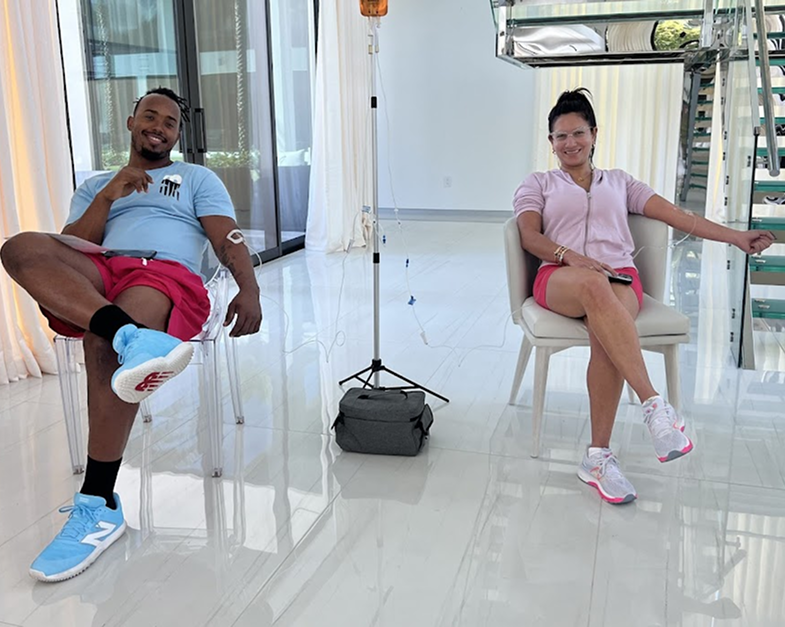 Two people receiving IV therapy, sitting indoors. Man in blue shirt, red shorts; woman in pink.