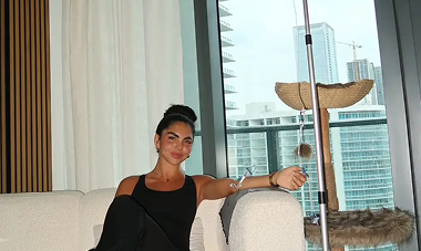 Woman in black outfit sits on a couch near a window, high-rise cityscape in the background.