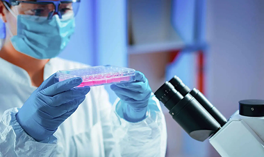 Scientist in lab coat, mask, and gloves holds a cell culture plate, near a microscope.
