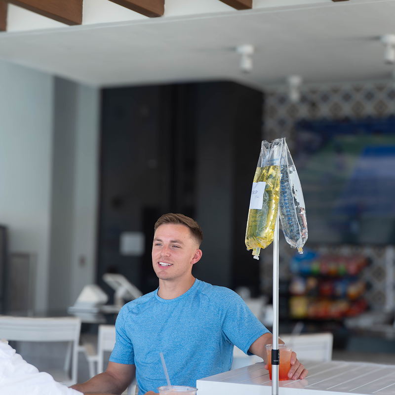 Man receiving IV drip while sitting at a bar. He is smiling, with orange and pink drinks nearby.
