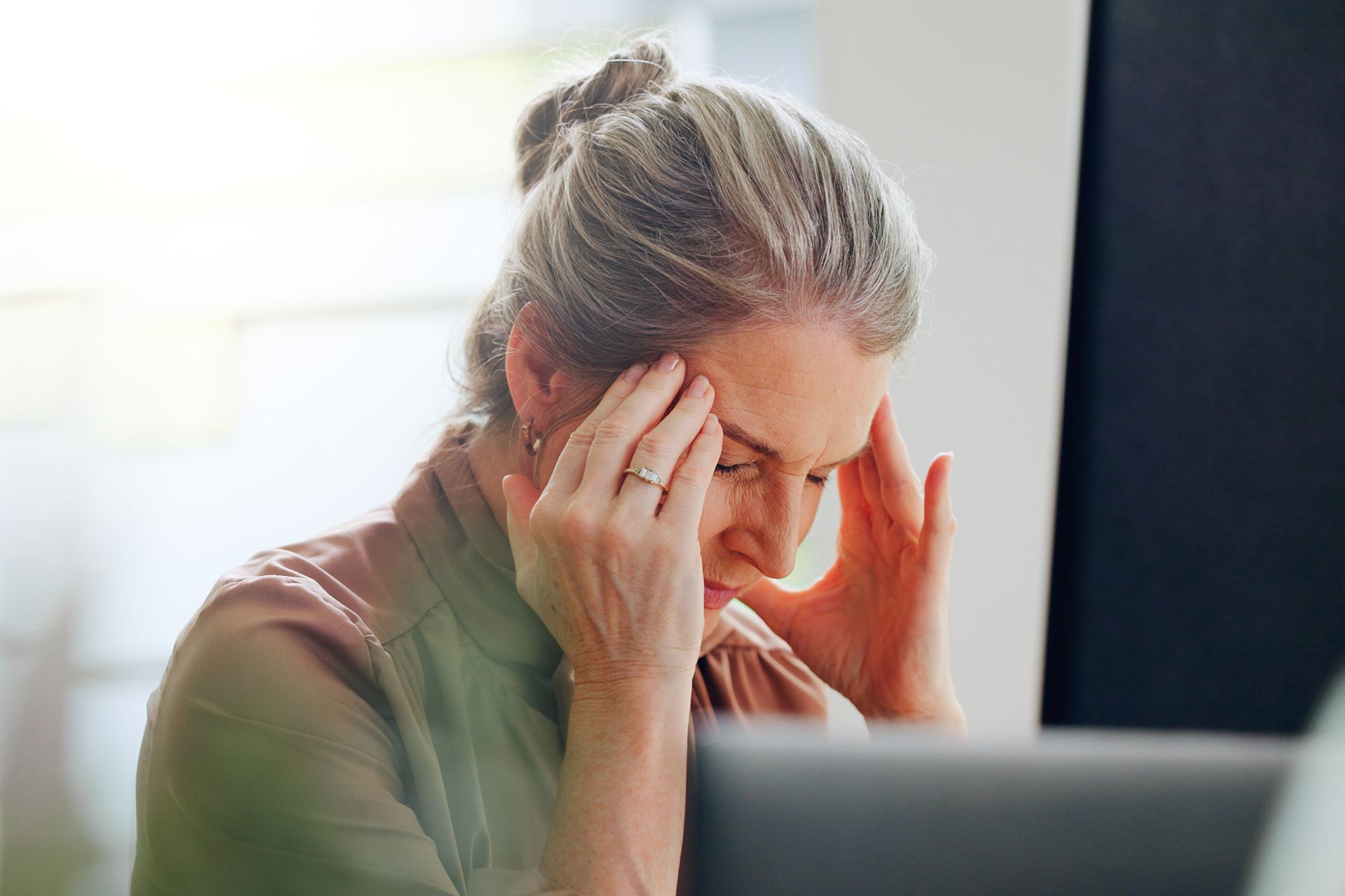 A person with gray hair sits at a laptop, holding their head with both hands in a gesture of stress or a headache.