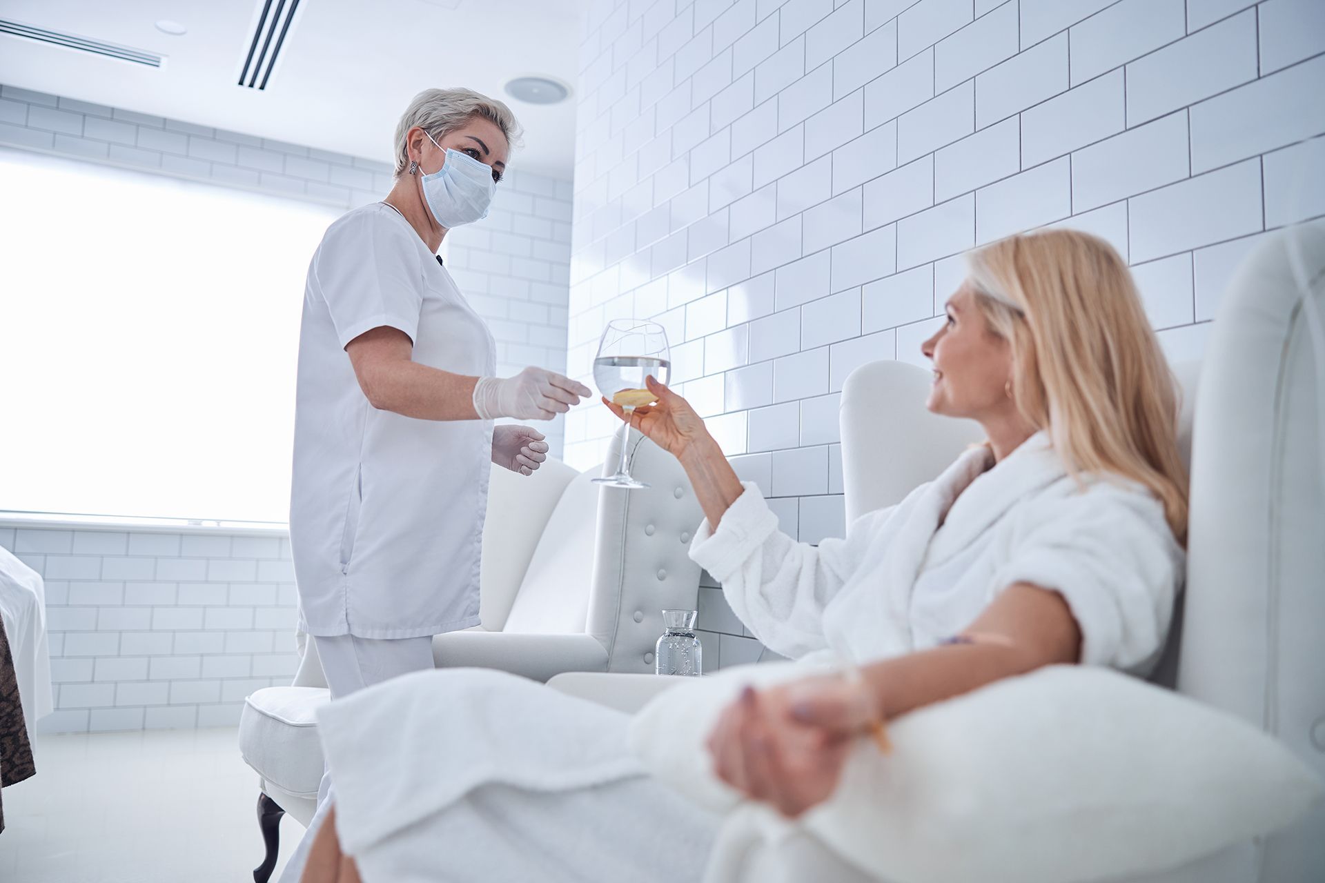 A person in a white uniform and mask hands a glass of water to another person in a white robe sitting in a spa setting.