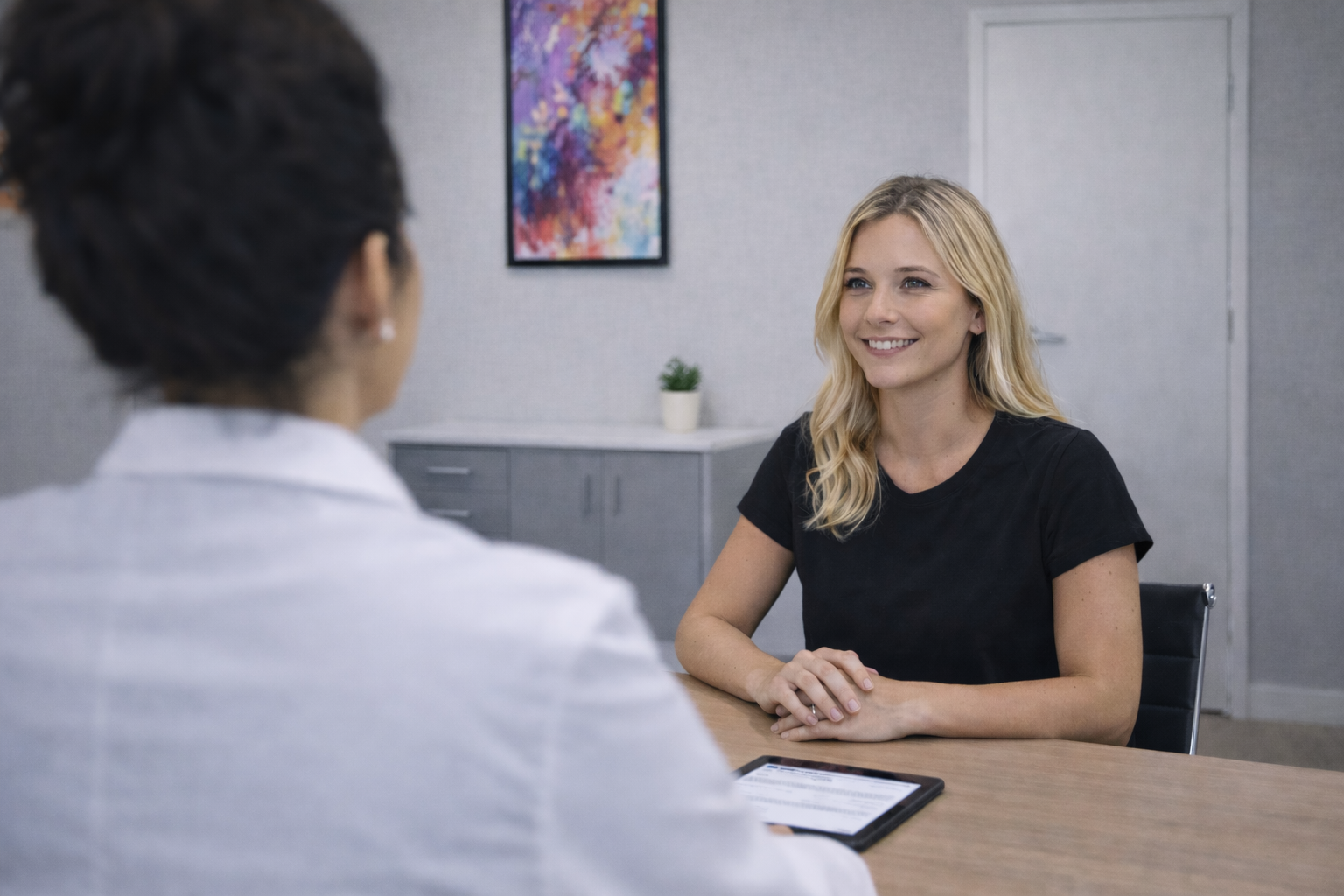 A person in a white lab coat sits across from a smiling person at a desk in an office with a painting on the wall.