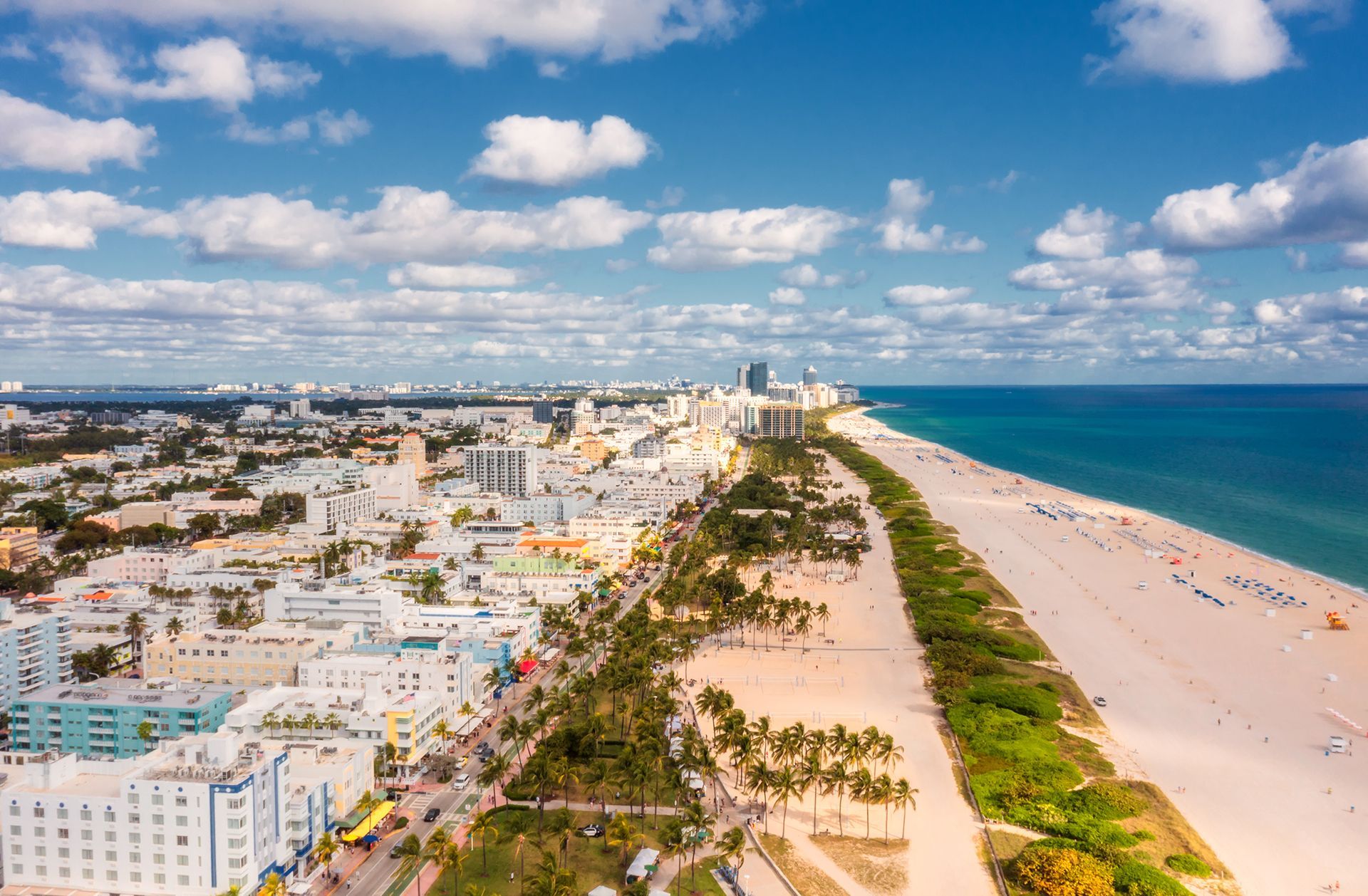 Aerial view of a sunny coastal city with white buildings, palm-lined streets, and a long sandy beach beside blue ocean.