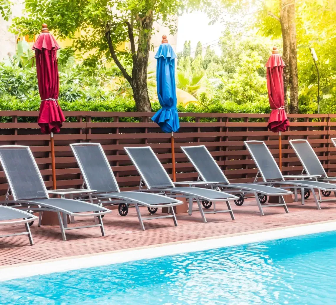 Poolside lounge chairs beside a turquoise swimming pool, with folded colorful umbrellas and trees in the background