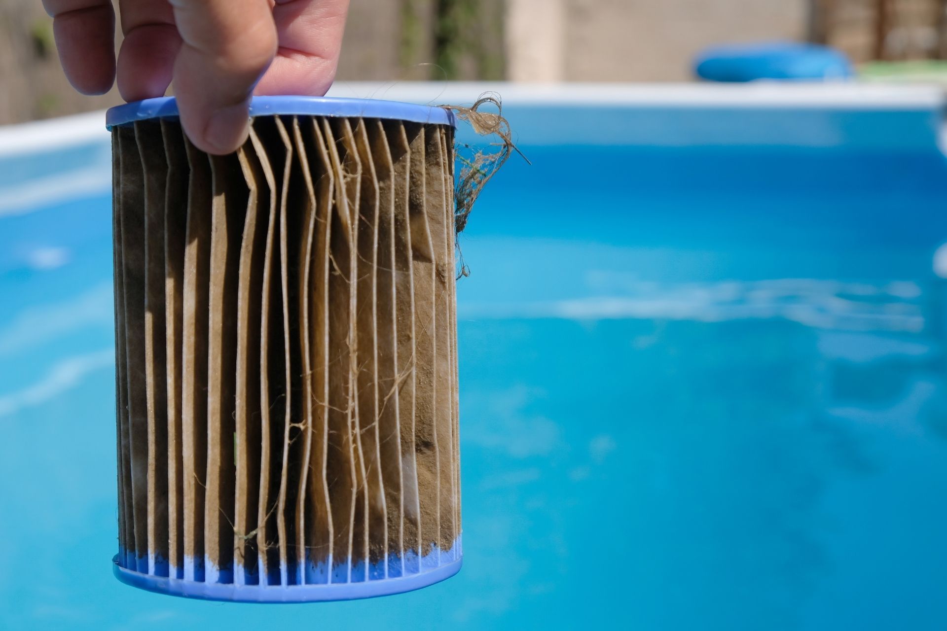 Hand holding a dirty pleated pool filter cartridge over blue pool water