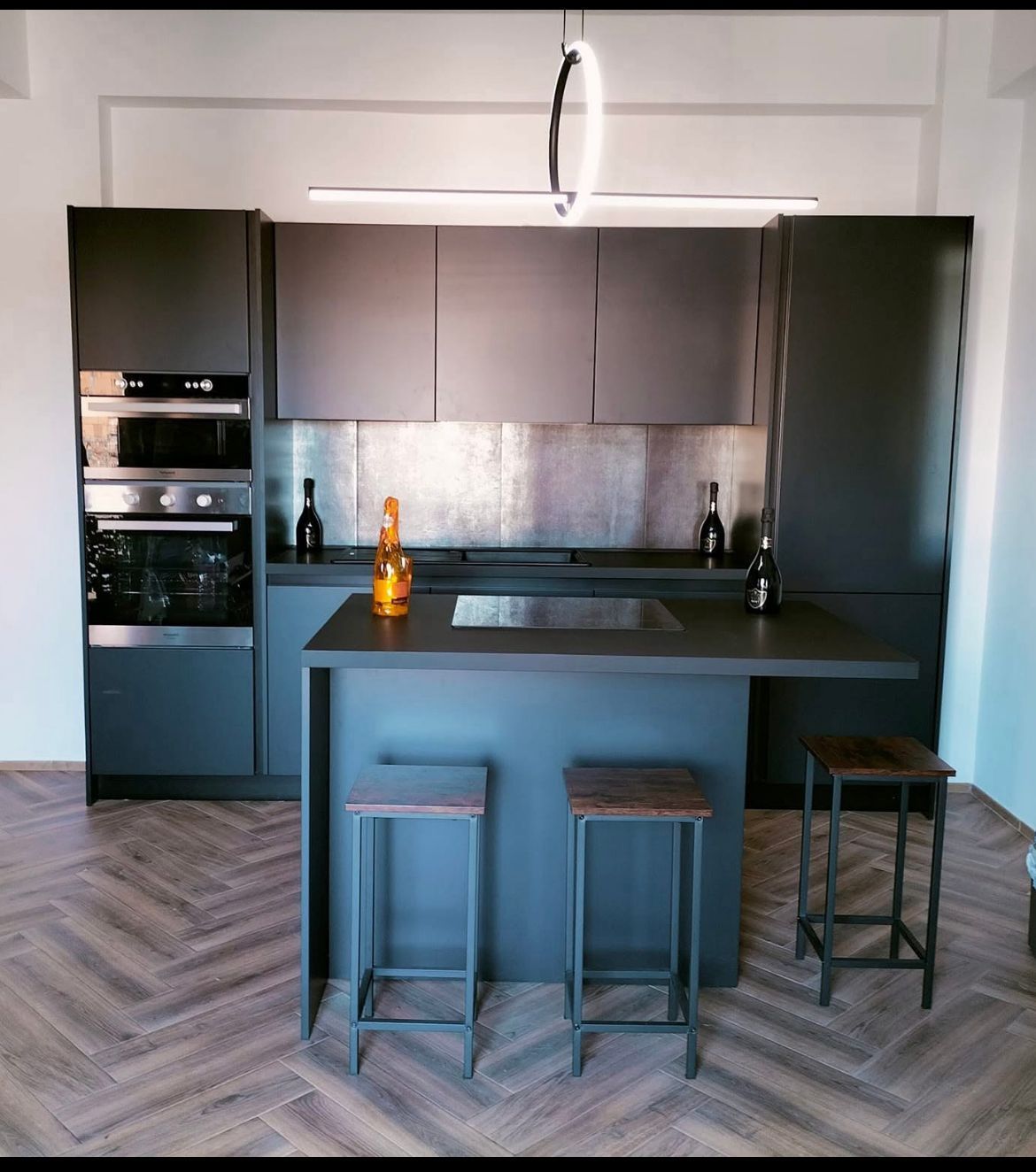 Una moderna cucina color grigio antracite con isola centrale, tre sgabelli da bar, pavimento in legno a spina di pesce e faretto ad anello.