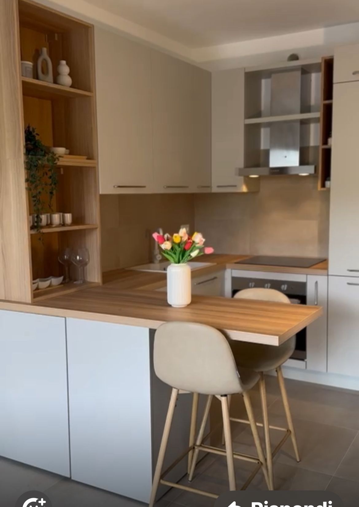 Una moderna cucina a L con mobili chiari, ripiani effetto legno, un'isola con due sgabelli e un vaso di fiori.