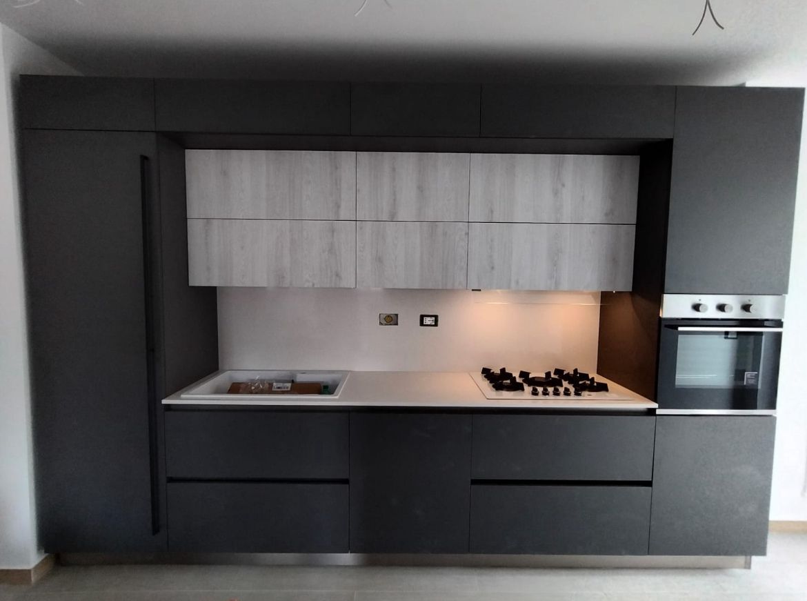 Una cucina moderna con mobili neri opachi, pensili in legno chiaro, piano di lavoro bianco e forno a incasso.