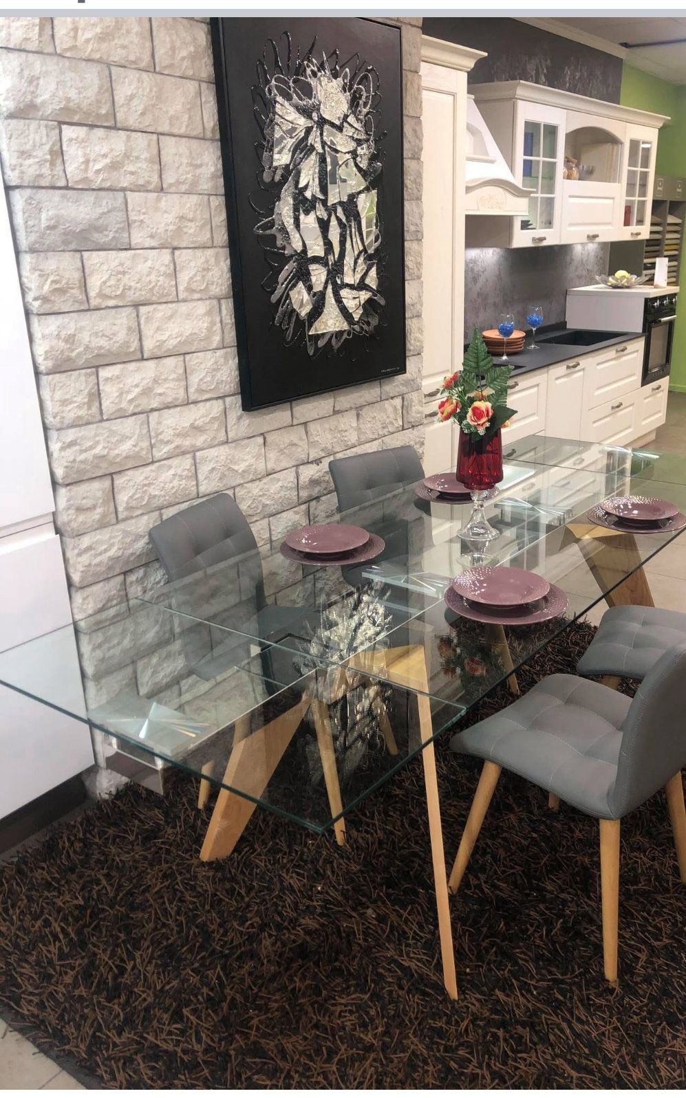 Un tavolo da pranzo con piano in vetro e sedie grigie, posizionato su un tappeto marrone di fronte a un muro di pietra con un grande dipinto astratto.