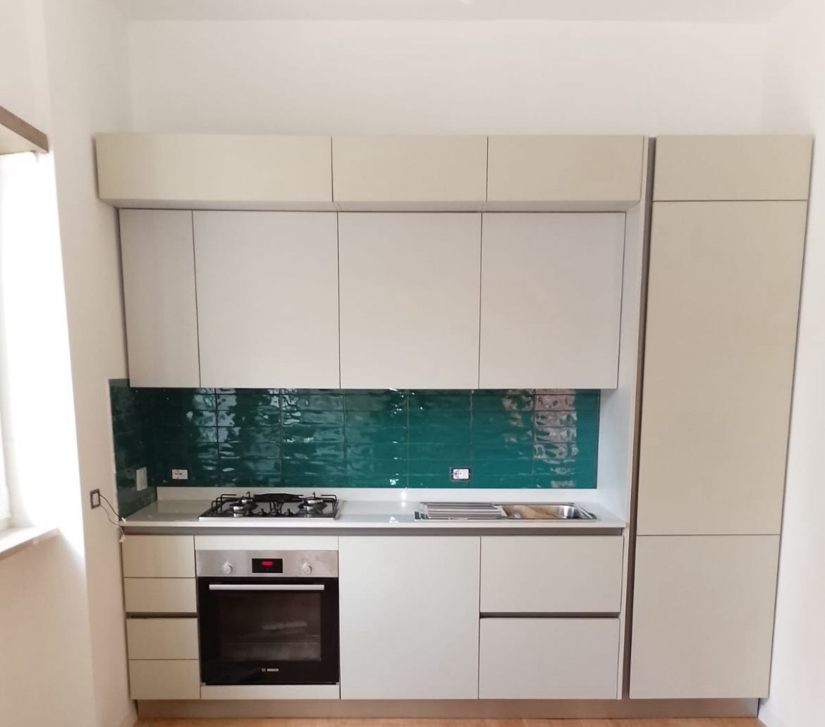 Una cucina moderna con mobili color beige chiaro opaco, forno in acciaio inox e paraschizzi rivestito con piastrelle color verde acqua lucido.