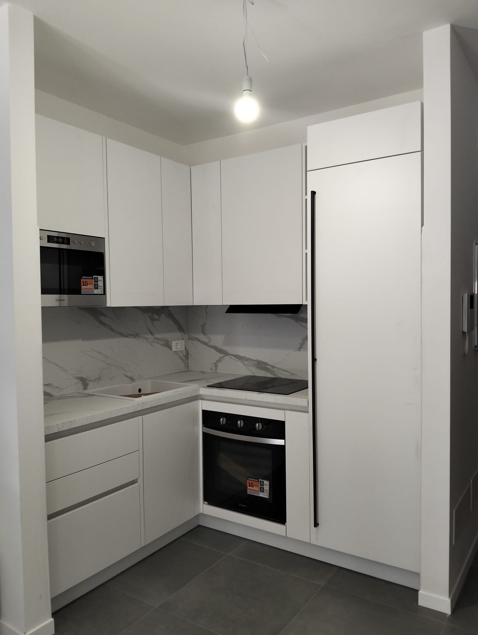 Una cucina moderna e compatta a forma di L, con mobili bianchi, forno a incasso, paraschizzi effetto marmo e pavimento grigio.