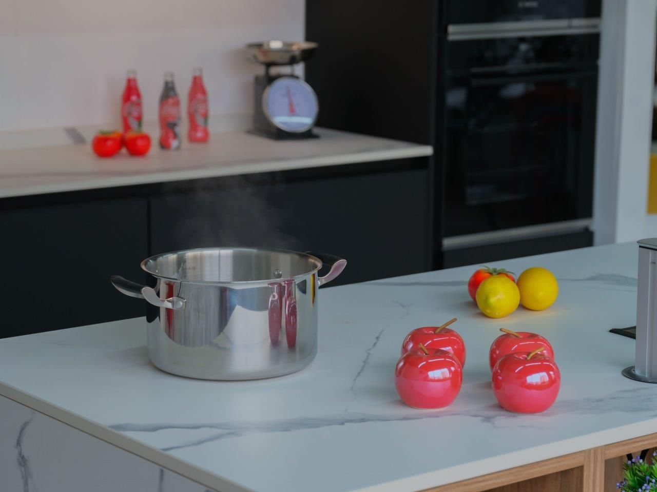 Una pentola fumante in acciaio inox è appoggiata su un'isola da cucina in marmo bianco, accanto a due mele rosse e due limoni.