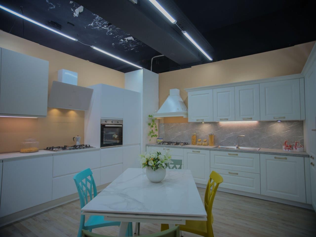 Una cucina moderna con mobili bianchi, un tavolo da pranzo centrale con una sedia color verde acqua e una gialla, e soffitto scuro.